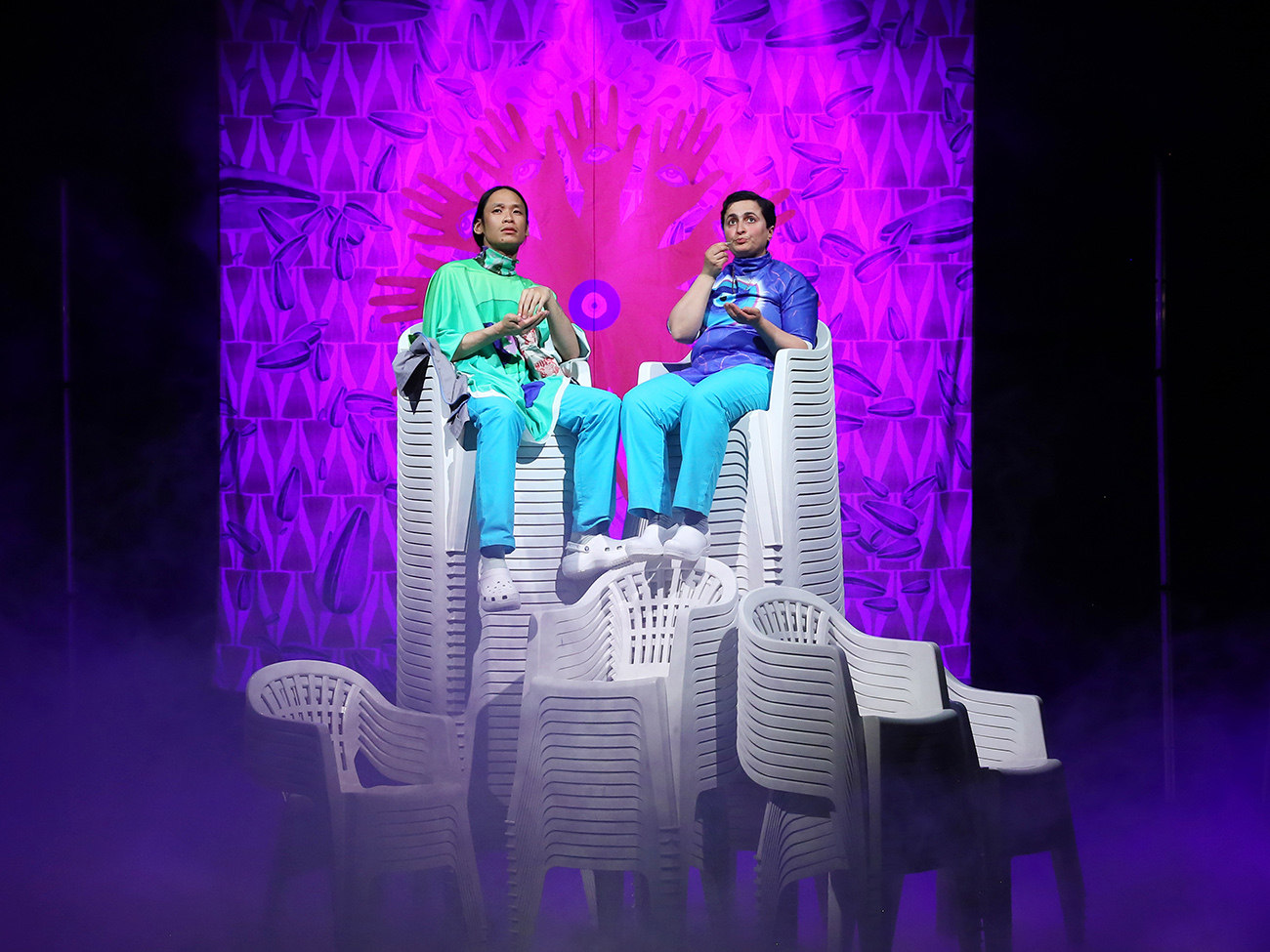 On stage, two people are sitting on a high pile of plastic chairs. Behind them is a bright purple curtain.