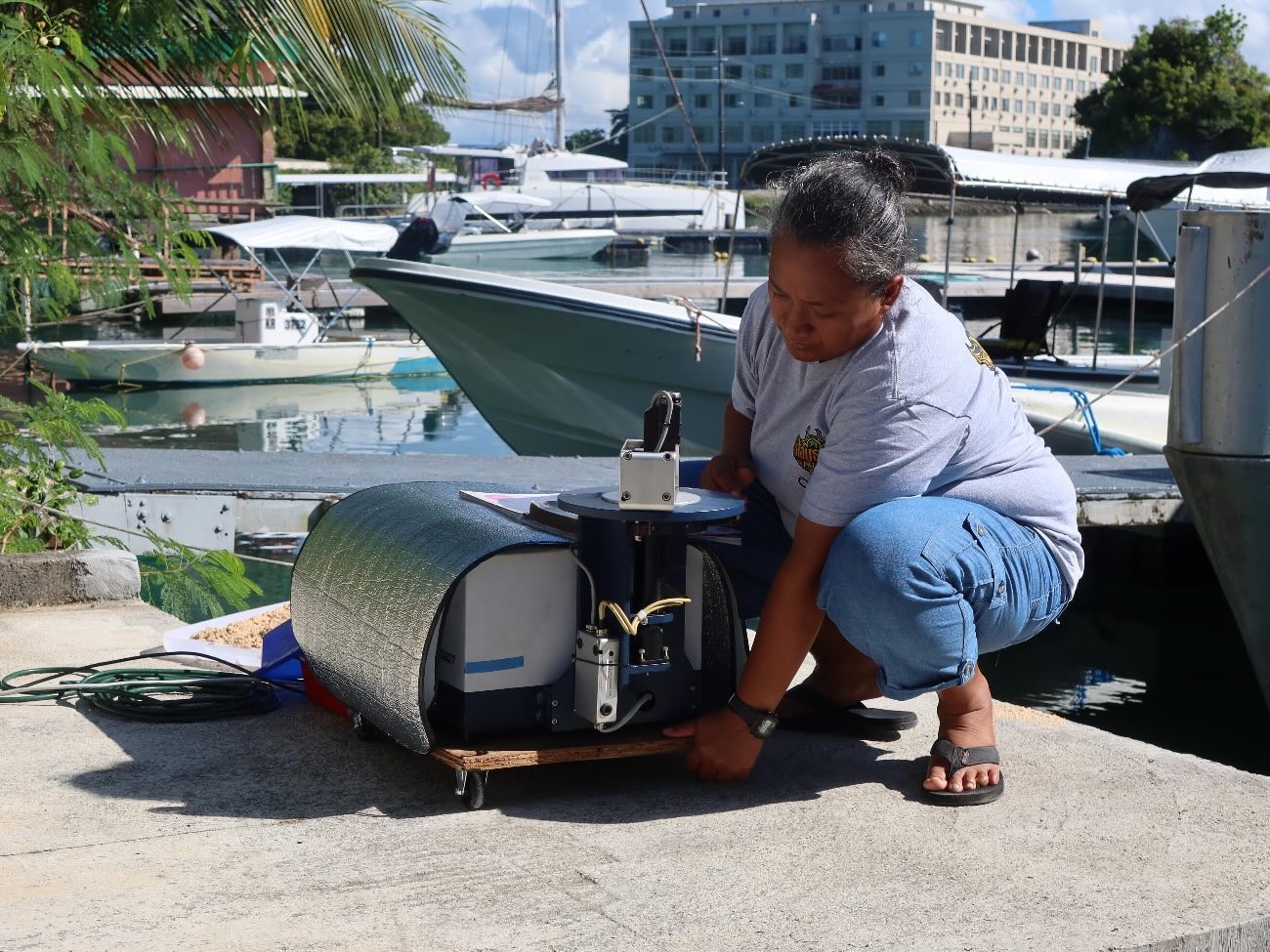 Sharon Patris is crouched next to a portable spectrometer against the backdrop of a harbor, taking measurements with it.