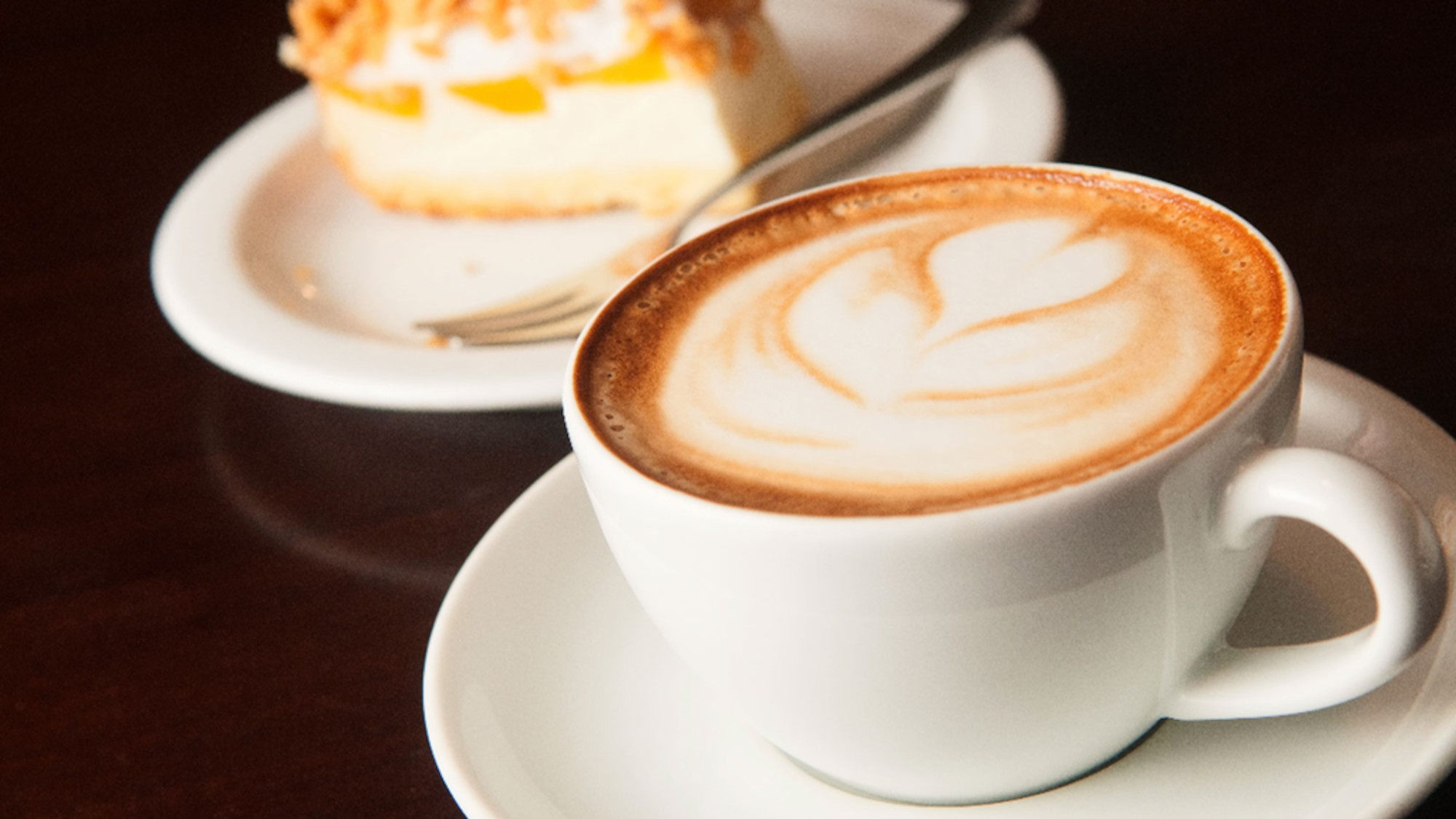 A cappuccino cup sits on a table with a slice of apple pie in the background